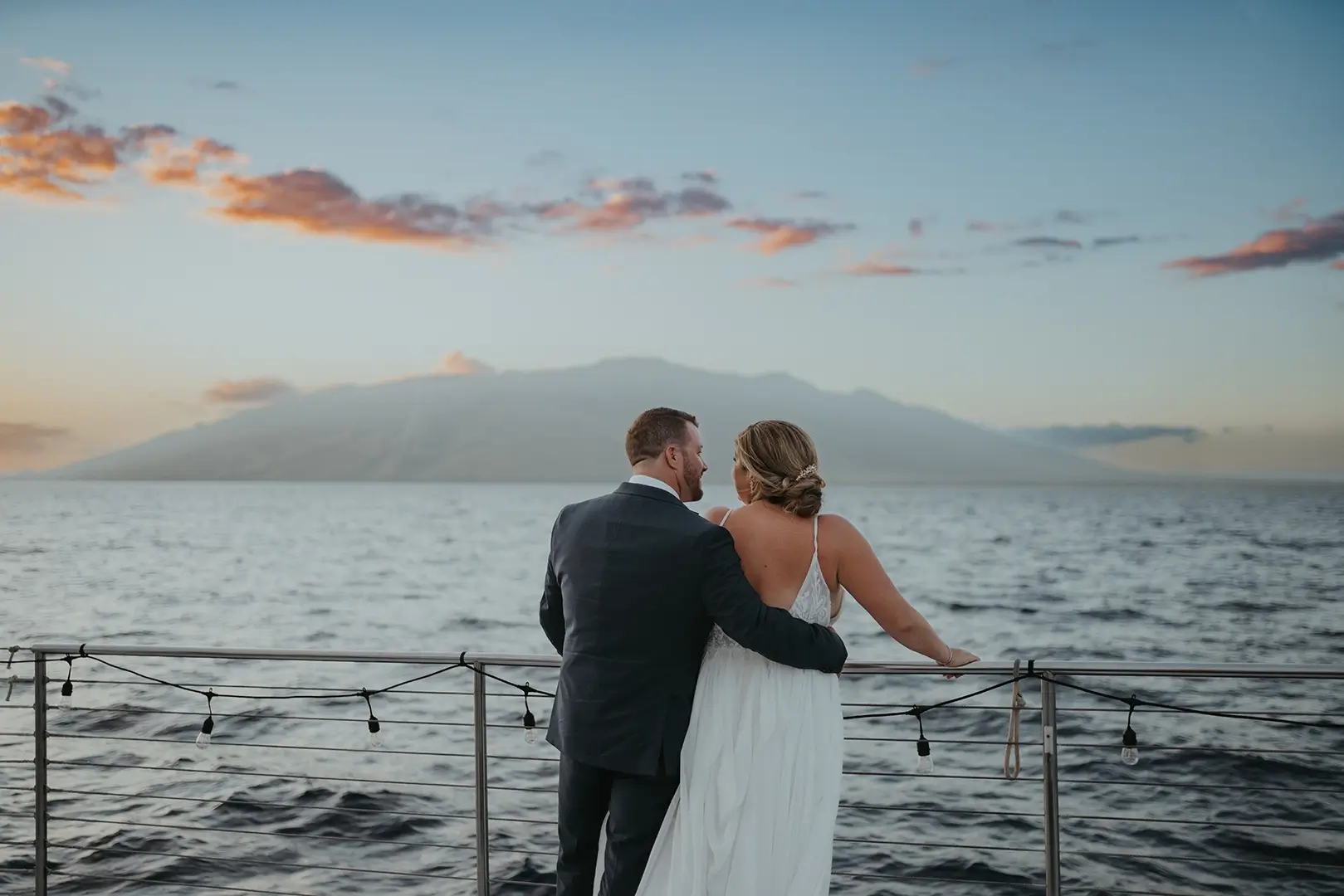 A couple stands on a dock, gazing at the serene ocean and distant mountains as the sun sets, casting a warm golden hue in the sky.