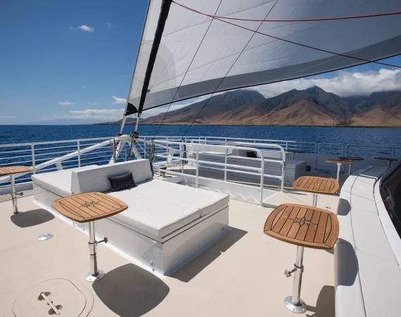 Wooden tables on a yacht deck overlook the ocean, with white railings and a blue sky dotted with clouds in the background.