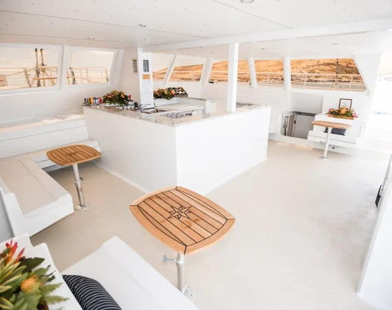 Wooden tables on a yacht deck overlook the ocean, with white railings and a blue sky dotted with clouds in the background.
