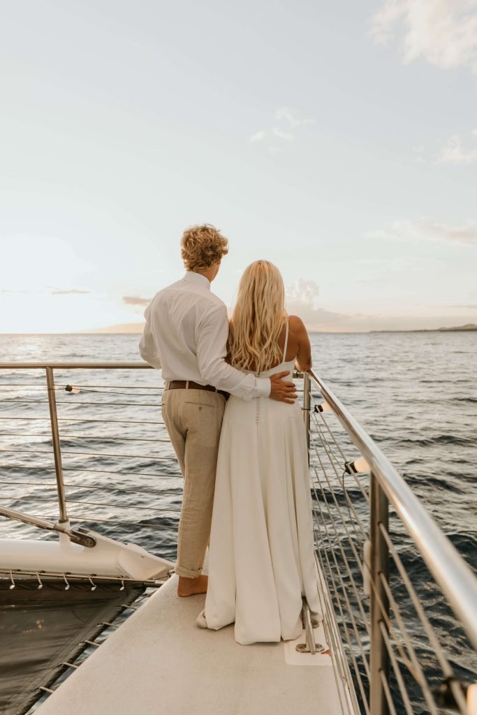 A couple stands on a boat, embracing and watching the sunset over the ocean, dressed in elegant attire.