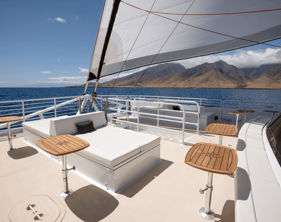 Sailing yacht deck featuring a sunbed and wooden tables, with scenic mountains and calm ocean in the background under clear blue skies.