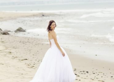A woman in a white wedding dress walks along a sandy beach with the ocean in the background under a cloudy sky.