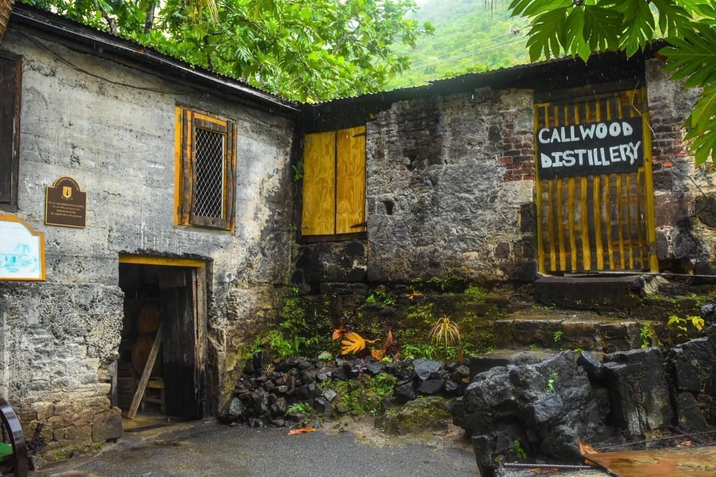 Exterior View Of Callwood Distillery, Showcasing Its Rustic Stone Walls And Partially Open Wooden Doors, Surrounded By Lush Green Foliage.