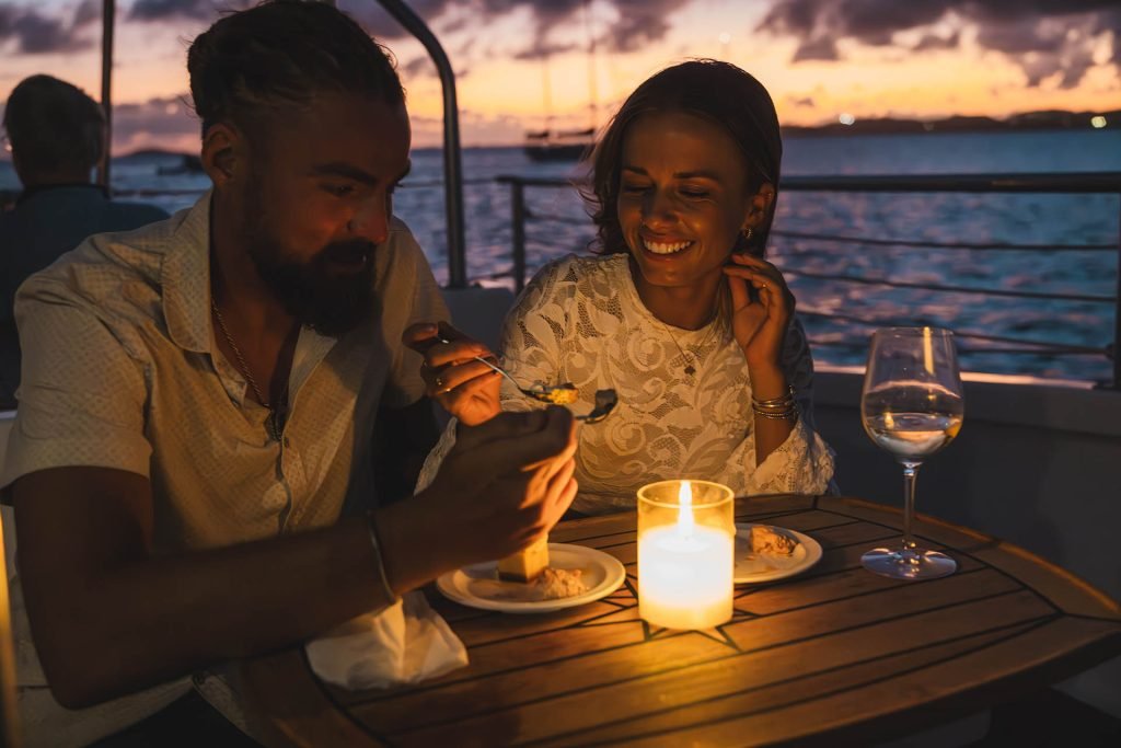 Couple Enjoying Sunset Sailing On A Catamaran While Having Dinner.