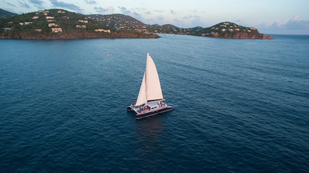 A Sailboat Cruising On Clear Blue Waters Near Hilly Islands Under A Lightly Cloudy Sky, Showcasing Cruz Bay Watersports.