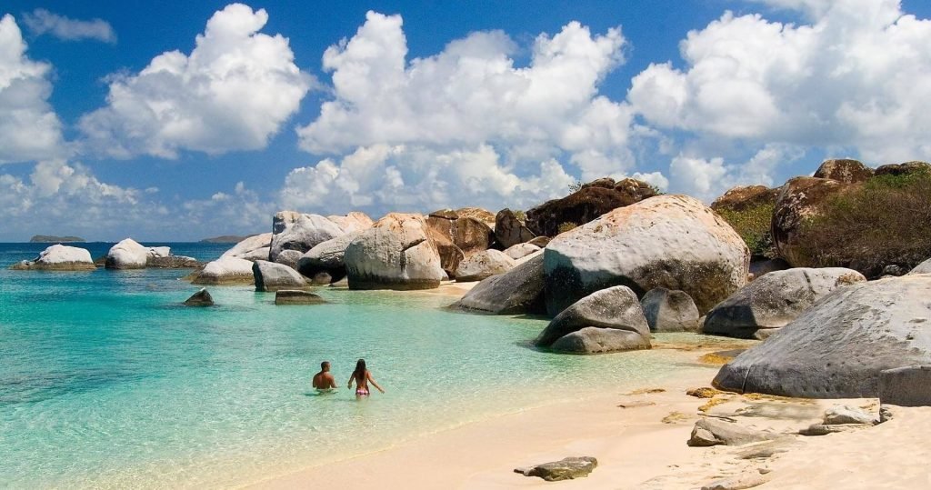 Scenic view of a tropical beach with turquoise waters, large granite boulders, and a clear blue sky, featuring a man and woman enjoying the shallow water at The Baths, Virgin Gorda.