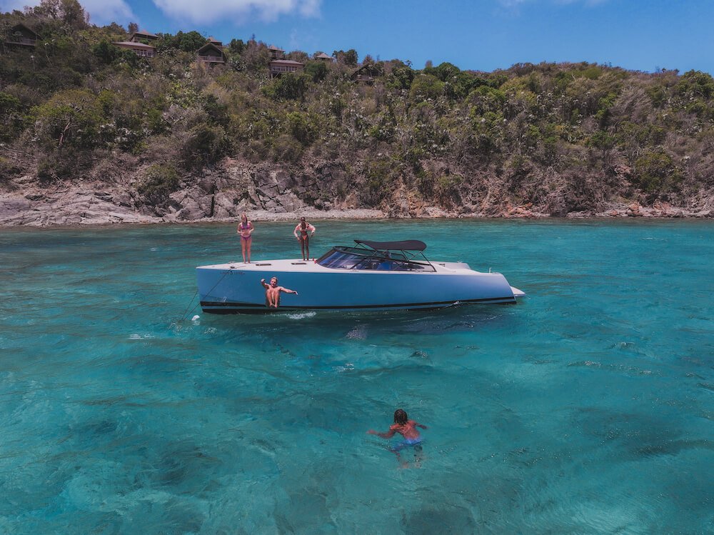 People enjoying a day on a yacht in clear blue water near a rocky shore with lush green vegetation.