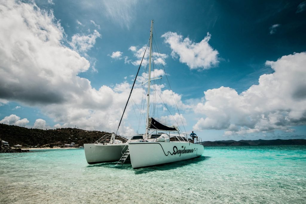 A catamaran named "Daydreamer" anchored in clear turquoise waters under a partly cloudy sky, with a scenic island in the background.