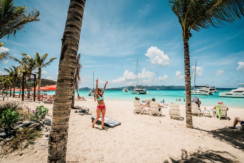 Woman celebrating on tropical beach with palm trees, white sand, turquoise water, and boats anchored offshore.