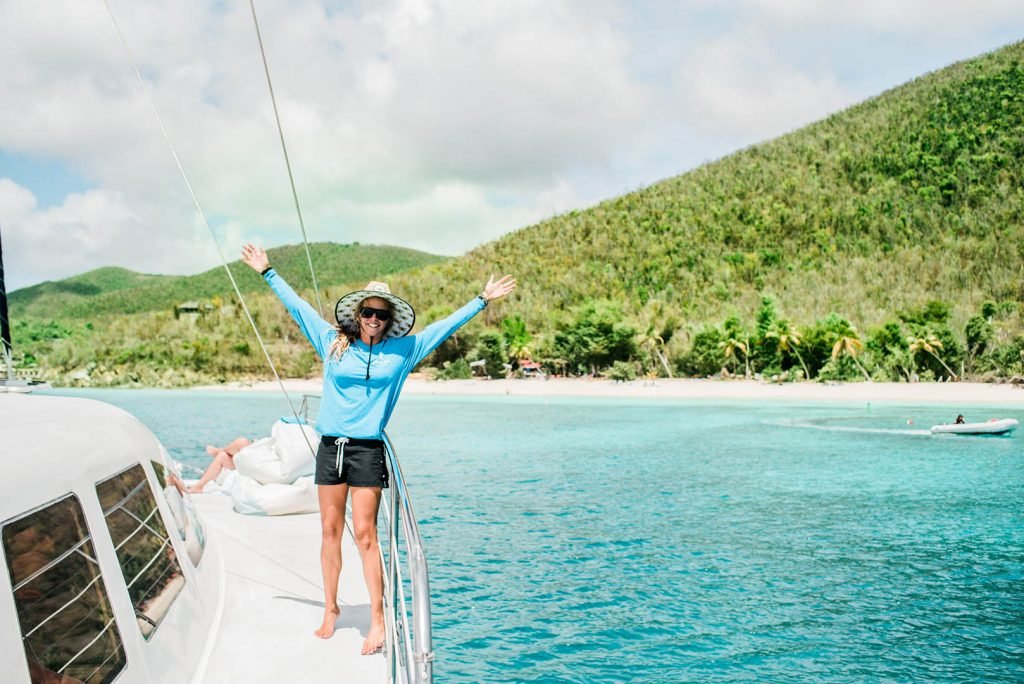 Woman In Sun Hat Celebrating With Arms Raised On Boat Deck Overlooking Turquoise Bay And Green Hills.