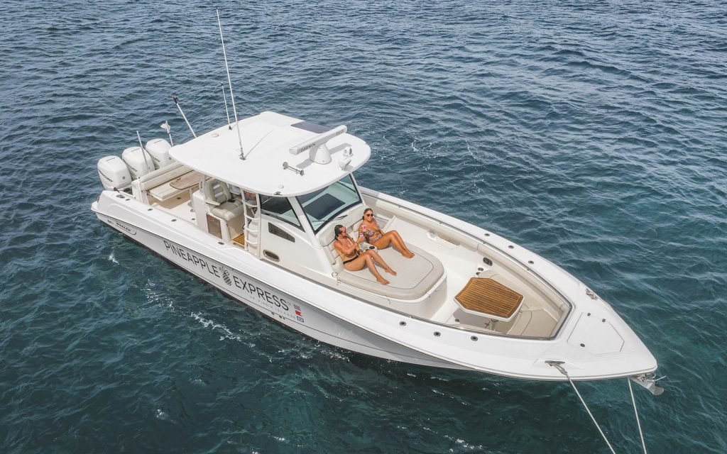A white boat named "PINEAPPLE EXPRESS" with two people lounging at sea.