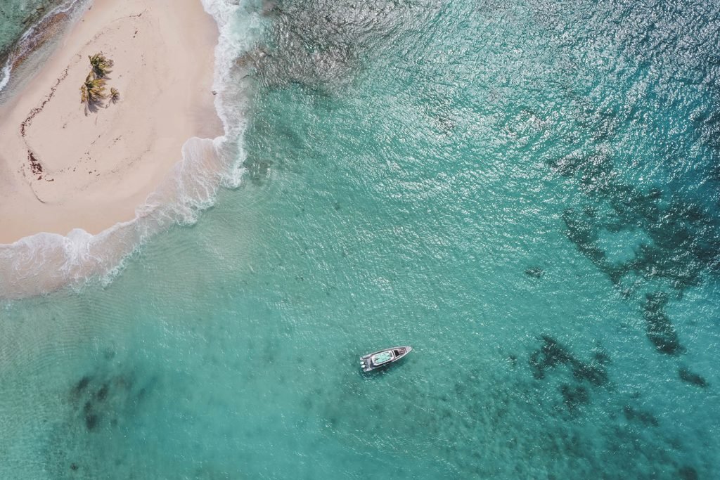 Aerial View Of A Small Boat Near A Tropical Island With Clear Blue Water And White Sand Beach.