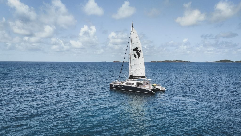 A catamaran with sails up on calm blue water, islands in the distance under a cloudy sky.