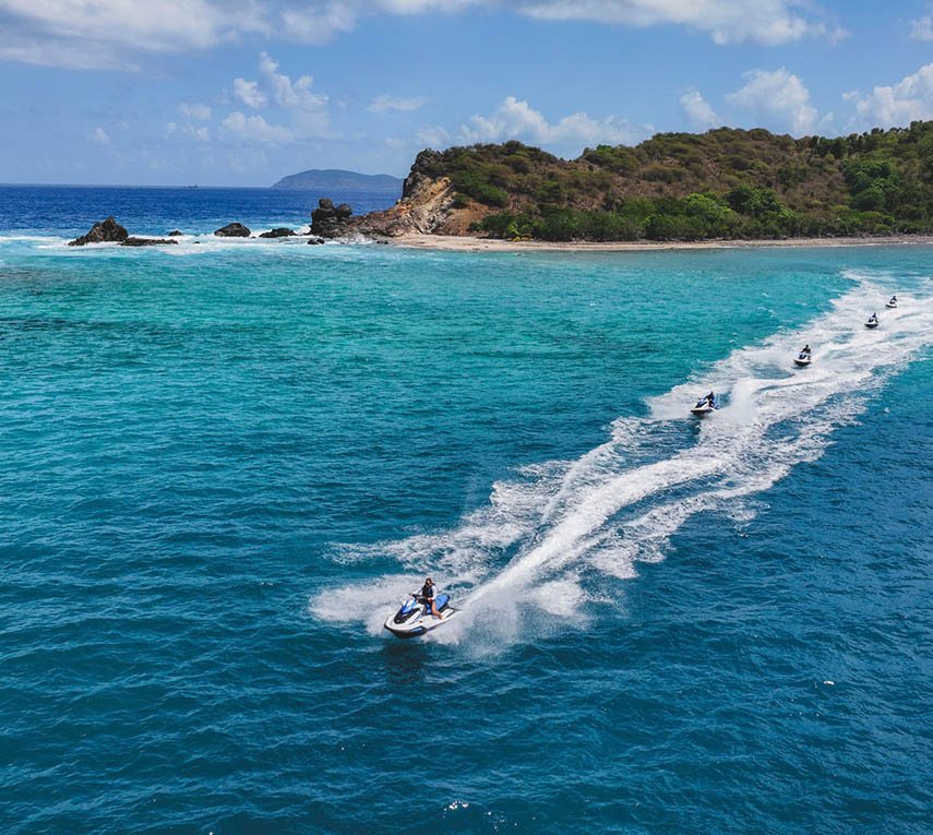 A Line Of Jet Skis Creating White Trails On A Turquoise Sea Near A Rocky Coast.
