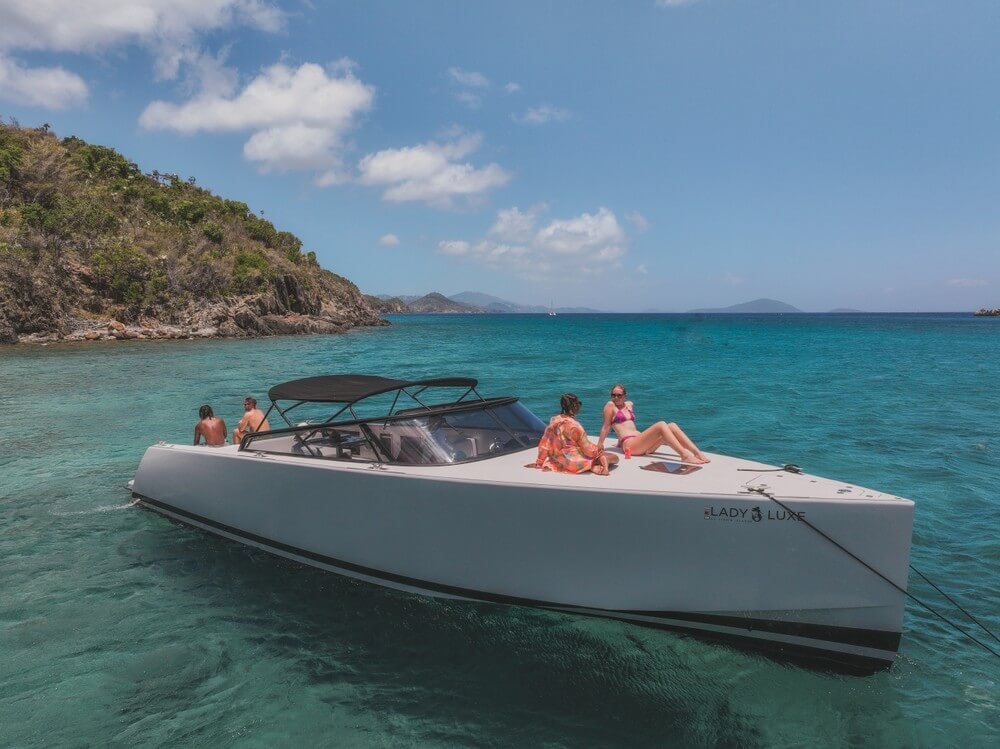 Luxury yacht anchored near a tropical island, with people relaxing on deck under a clear blue sky.