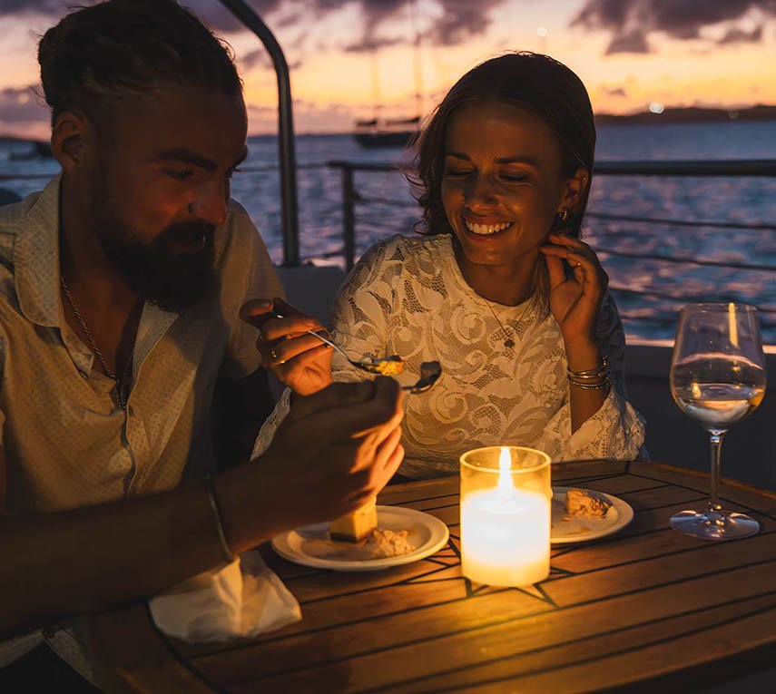Two people enjoying a candlelit dinner on a boat at sunset.