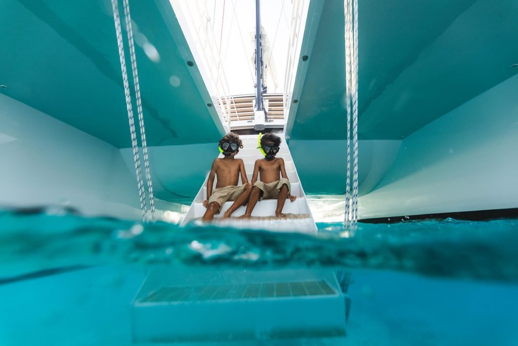 Two Children Sitting On A Boat'S Steps Wearing Snorkeling Masks, Ready To Swim In Clear Blue Water.