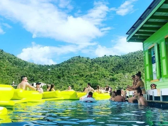 Group of people enjoying a sunny day at a pool with inflatable yellow floats, surrounded by lush green hills and a bright blue sky.