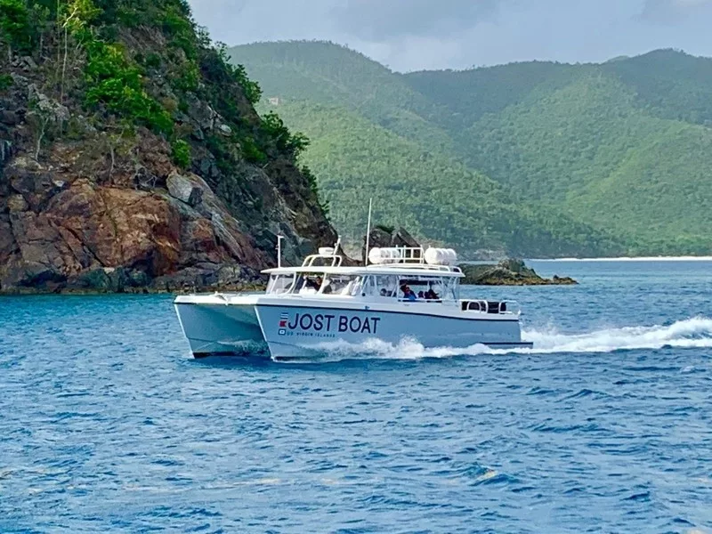 A boat named "Jost Boat" cruising through clear blue waters with lush green hills in the background.