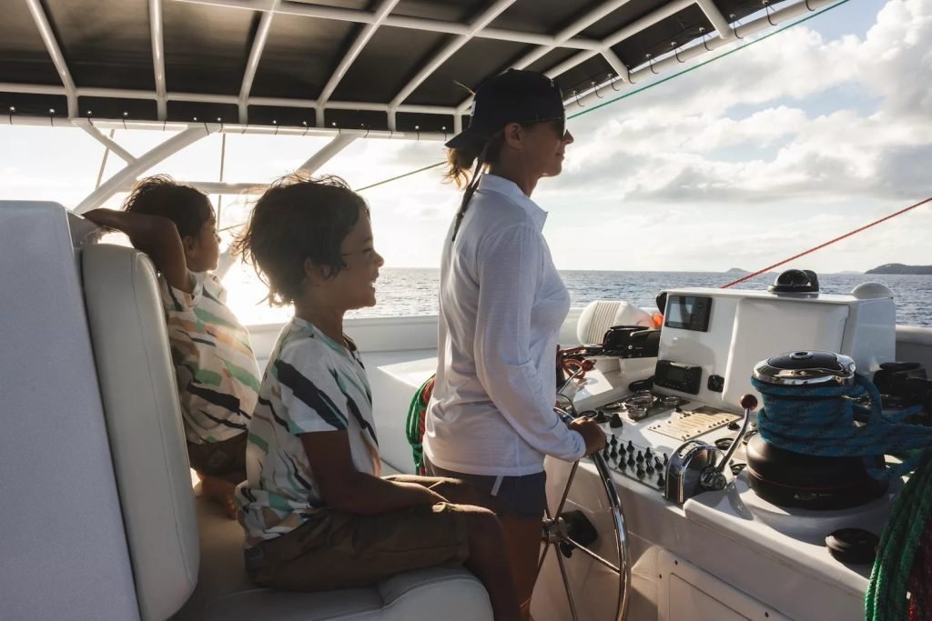 Family enjoying a boat ride, woman steering and children sitting beside her, scenic ocean view.