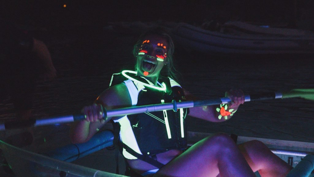 Person enjoying night kayaking with neon face paint and glow stick accessories.